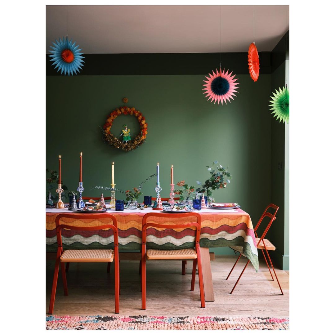 Festive dining room with a table set for a party, featuring colorful tablecloth, candles, decorations, chairs, and green walls.