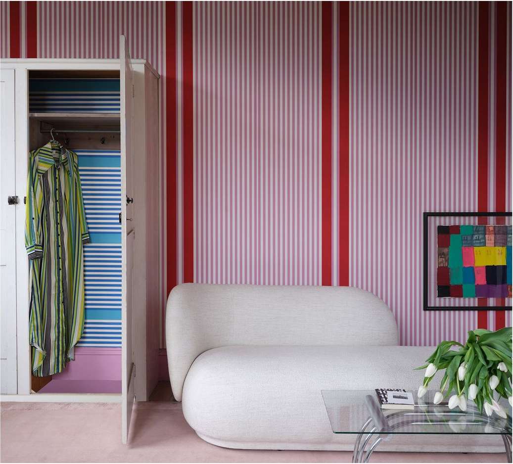 Interior featuring a cream couch, glass coffee table with books, a wardrobe, flowers, and striped pink wallpaper.