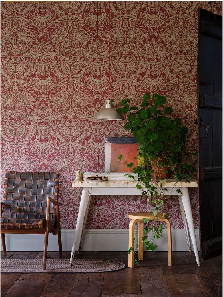 Cozy interior with a patterned red and beige wallpaper, woven chair, a small table, and a stool in a warm-toned room.