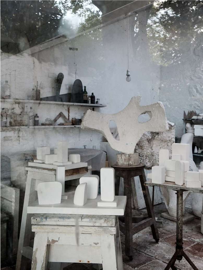 Indoor artist studio with abstract sculptures and geometric white plaster shapes on tables, illuminated by natural light streaming through windows.