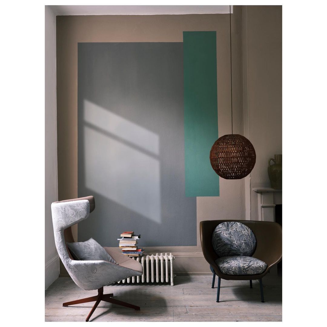 Modern interior with armchairs, woven pendant light, books, and radiator against a wall painted with geometric shapes.