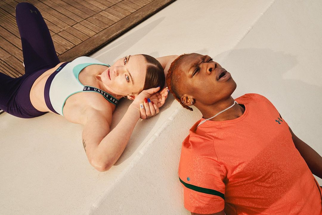 Two athletic women in sportswear, one with blue and white top, the other in coral top, lying together in studio for Under Armour.