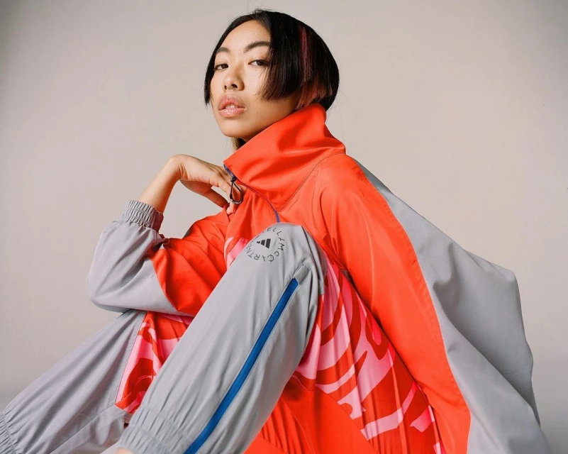 Asian woman in neon coral and grey Adidas tracksuit poses in studio. Apparel features Stella McCartney logo. Beauty shot, fashion editorial.