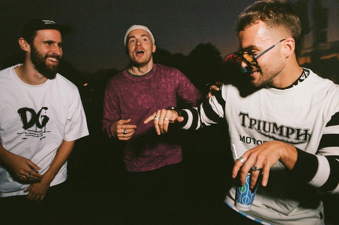 Three young adult men stand outside at dusk, talking. One wears sunglasses and holds a blue paper cup with a straw. T-shirts are branded.