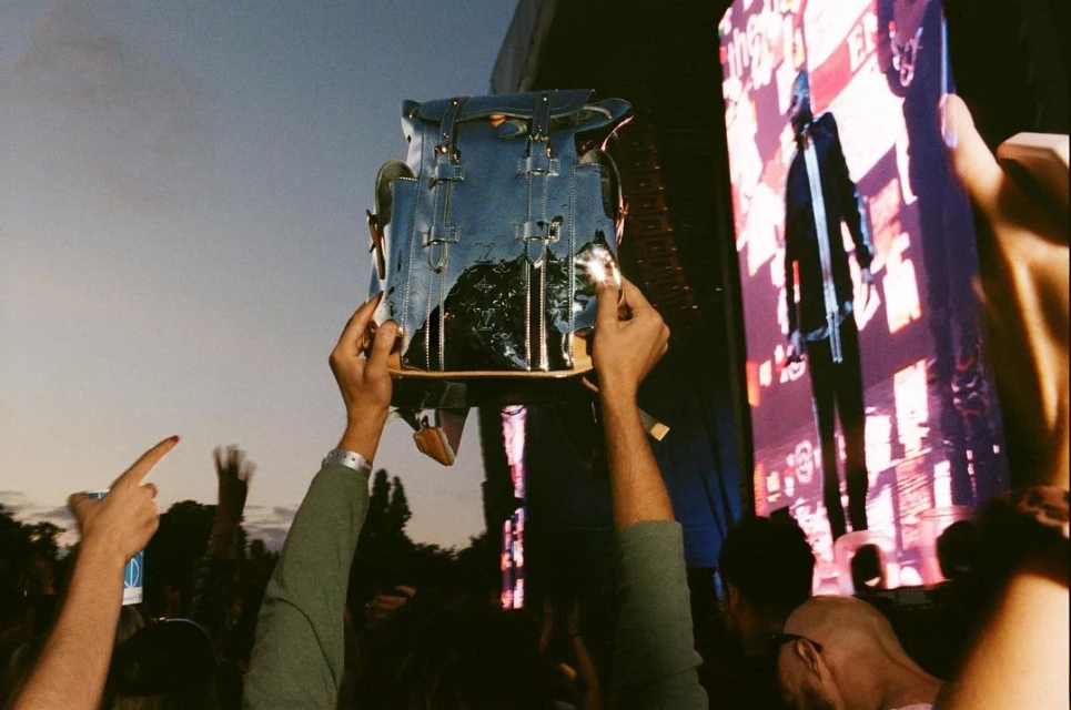At a concert, arms raise a Louis Vuitton backpack against a stage backdrop with a performer and a crowd of people visible.