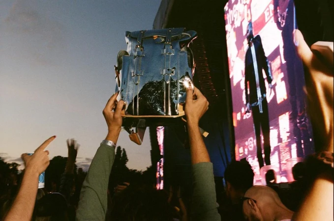 At a concert, arms raise a Louis Vuitton backpack against a stage backdrop with a performer and a crowd of people visible.