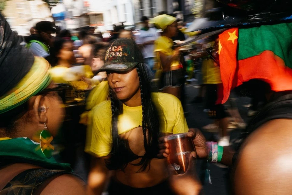 Street scene captures a woman in a yellow shirt and cap, holding a cup amidst a vibrant crowd with flags and movement.