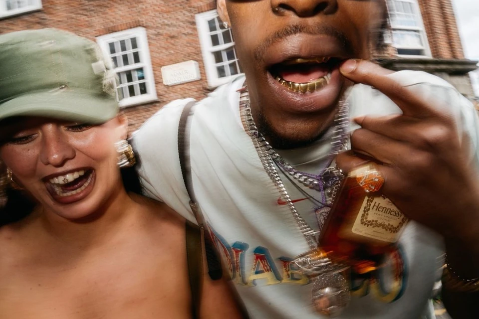 Two people with open mouths, gold teeth, and jewelry. One person holds a Hennessy bottle outside with a blurred brick building background.