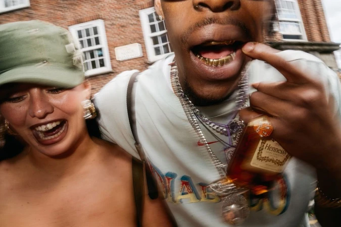 Two people with open mouths, gold teeth, and jewelry. One person holds a Hennessy bottle outside with a blurred brick building background.