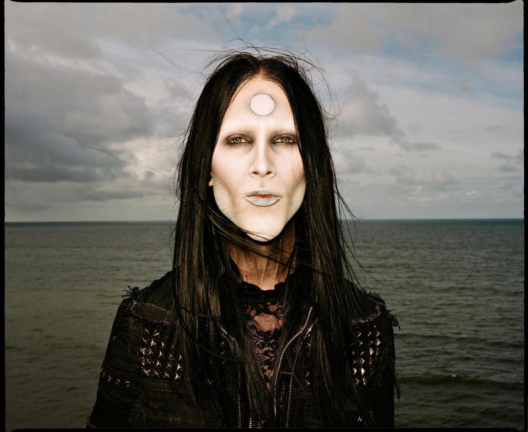 Person with white makeup and forehead dot standing in front of a cloudy sky and ocean, wearing a black studded jacket and black hair.