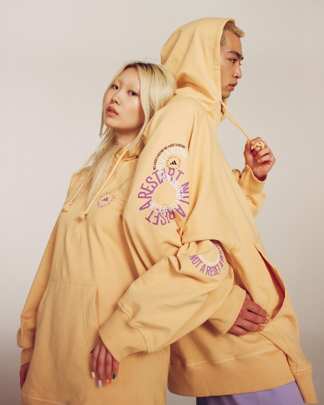 Two models pose back-to-back in matching apricot hoodies with graphic text, displaying casual fashion against a neutral backdrop.