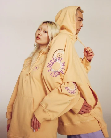 Two models pose back-to-back in matching apricot hoodies with graphic text, displaying casual fashion against a neutral backdrop.