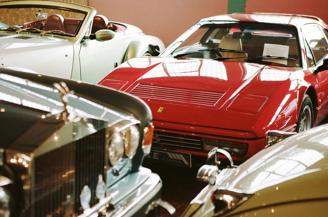 Several classic cars in a showroom featuring a red Ferrari, a white convertible, and a dark vintage Rolls Royce in an indoor setting.