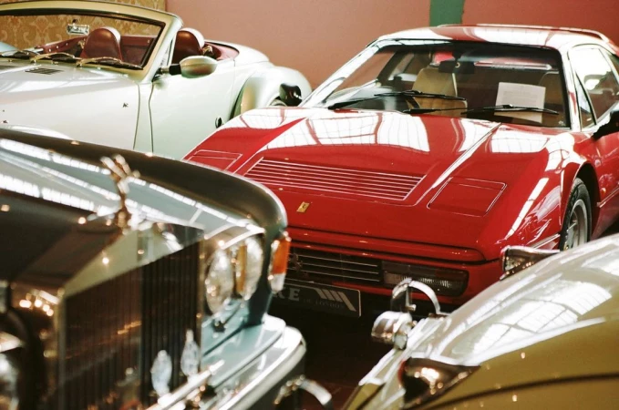 Several classic cars in a showroom featuring a red Ferrari, a white convertible, and a dark vintage Rolls Royce in an indoor setting.