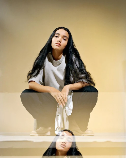 Portrait of a young adult woman with long dark hair in a crouched pose wearing casual clothes, set against a soft yellow background in a studio.