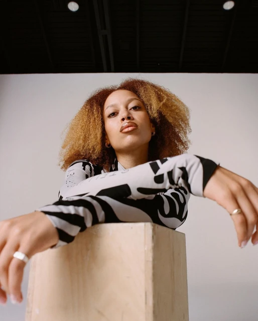 A young woman with curly hair poses confidently indoors, leaning against a light wooden cube in a casual black and white patterned top.