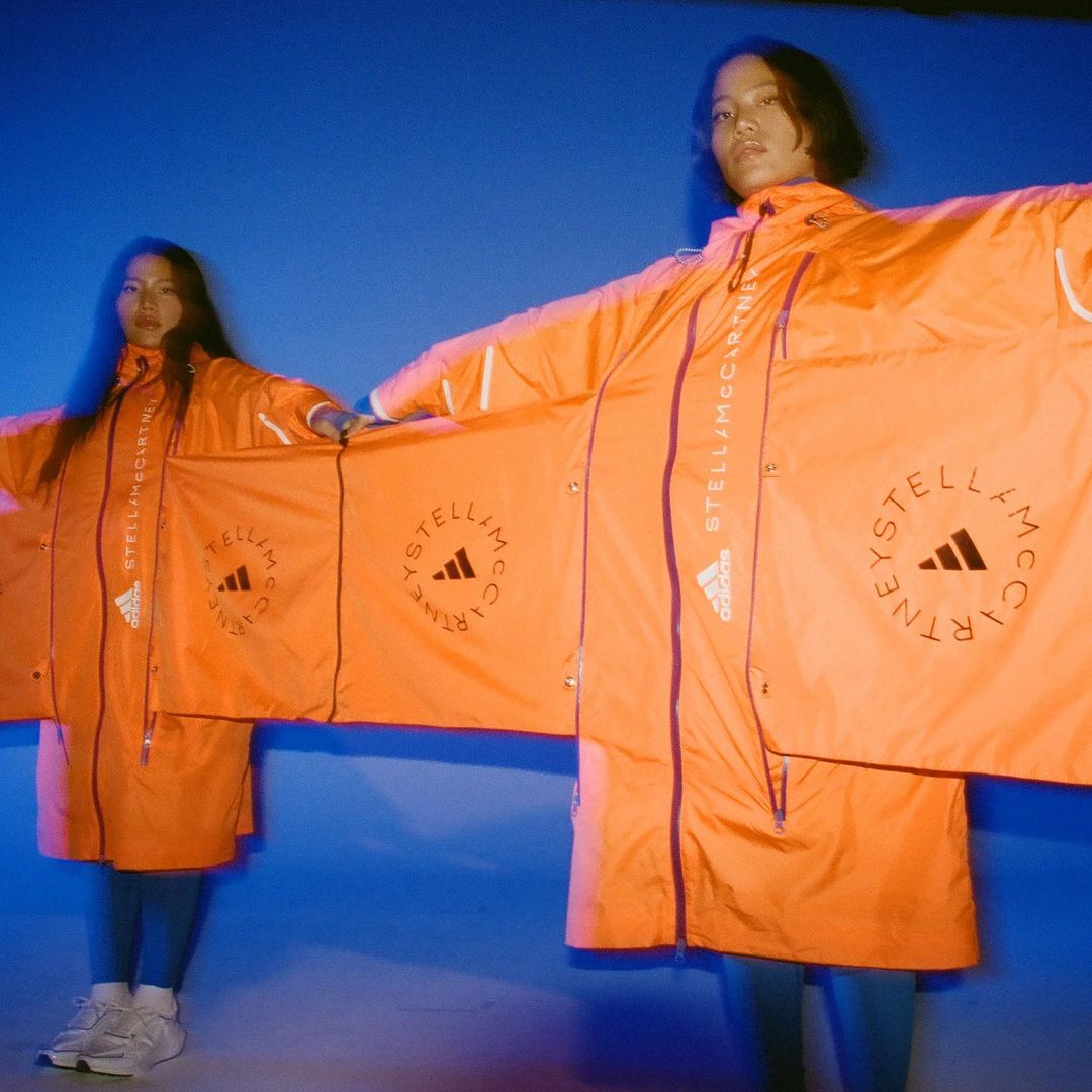 Two Asian women pose in matching orange Stella McCartney x Adidas logo raincoats against a blue backdrop in a fashion forward shot.