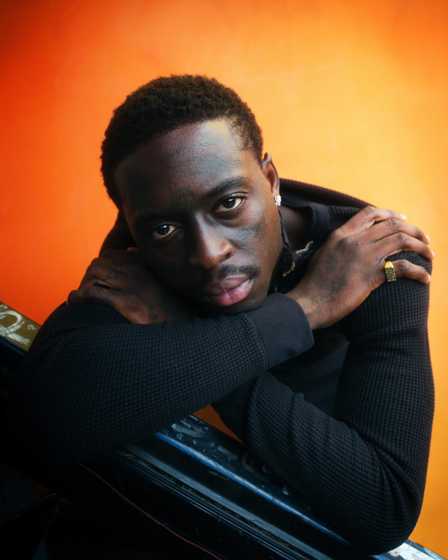 Portrait of a young Black man with a thoughtful expression, resting his arms on a dark surface against an orange backdrop.