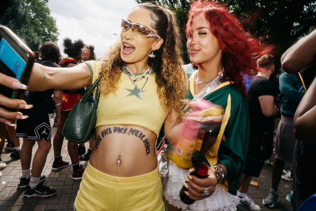 Two young women pose in trendy summer outfits at an outdoor event, taking a selfie in a crowd, showcasing fashion and youth culture.