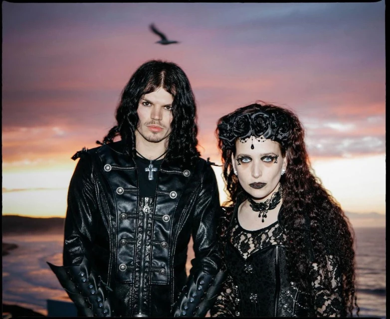 A gothic couple in dark clothing poses outdoors at dusk near the ocean, featuring intricate black attire and dramatic makeup.