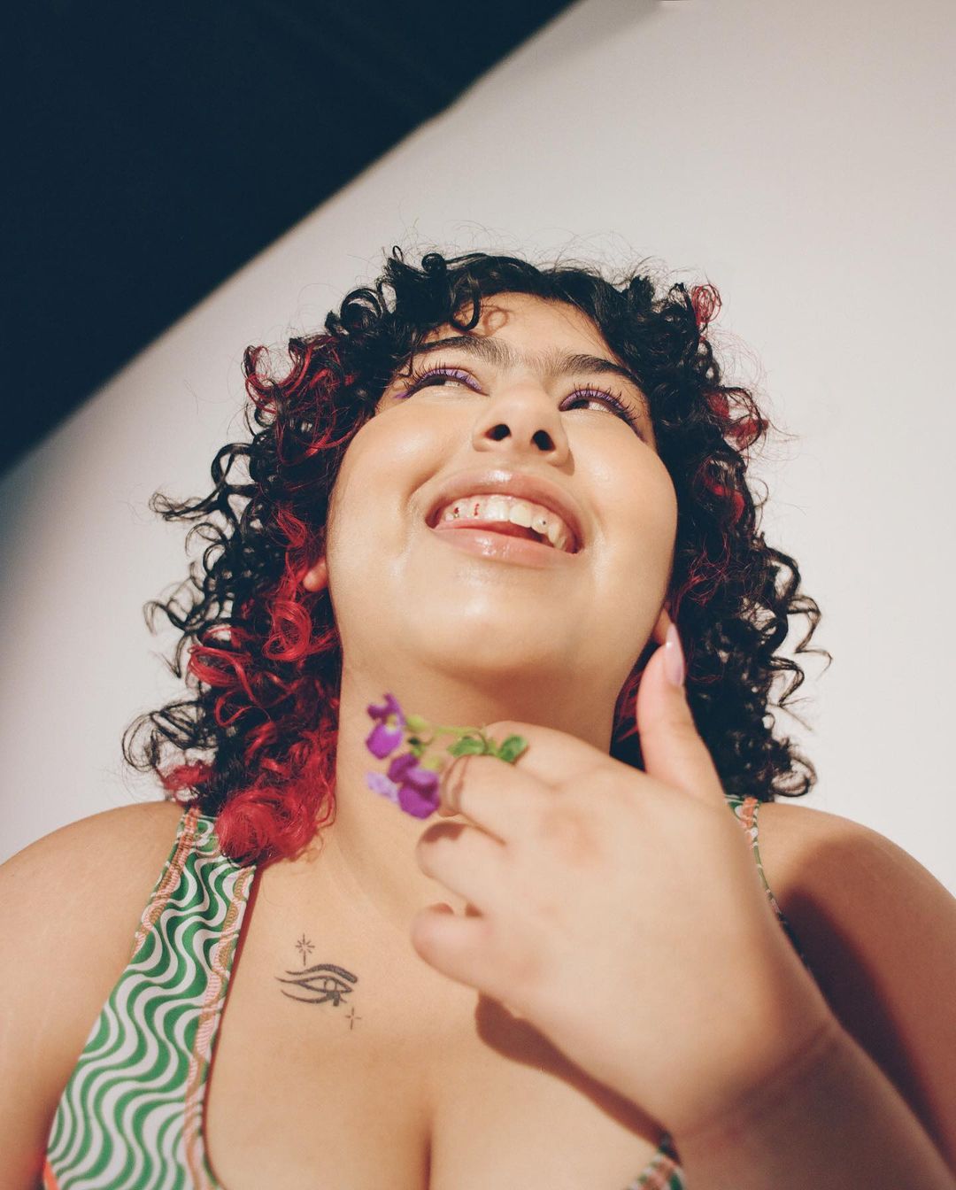 Smiling woman with curly red-highlighted hair holds purple flowers against a white backdrop, looking up with a tattoo visible on her chest.
