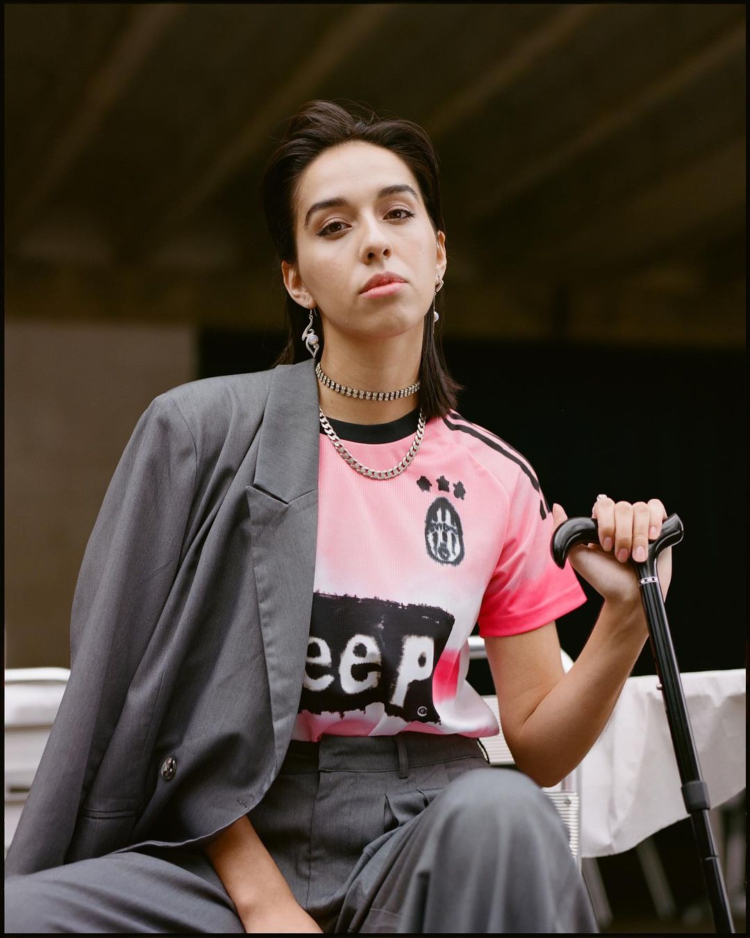Woman in soccer jersey and blazer posing with cane, gray pants, and chain necklaces in an urban setting, medium shot, fashion editorial.