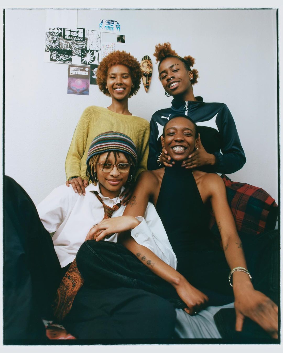 Portrait of four young women with diverse hairstyles smiling indoors against a white wall with posters and decorative mask.