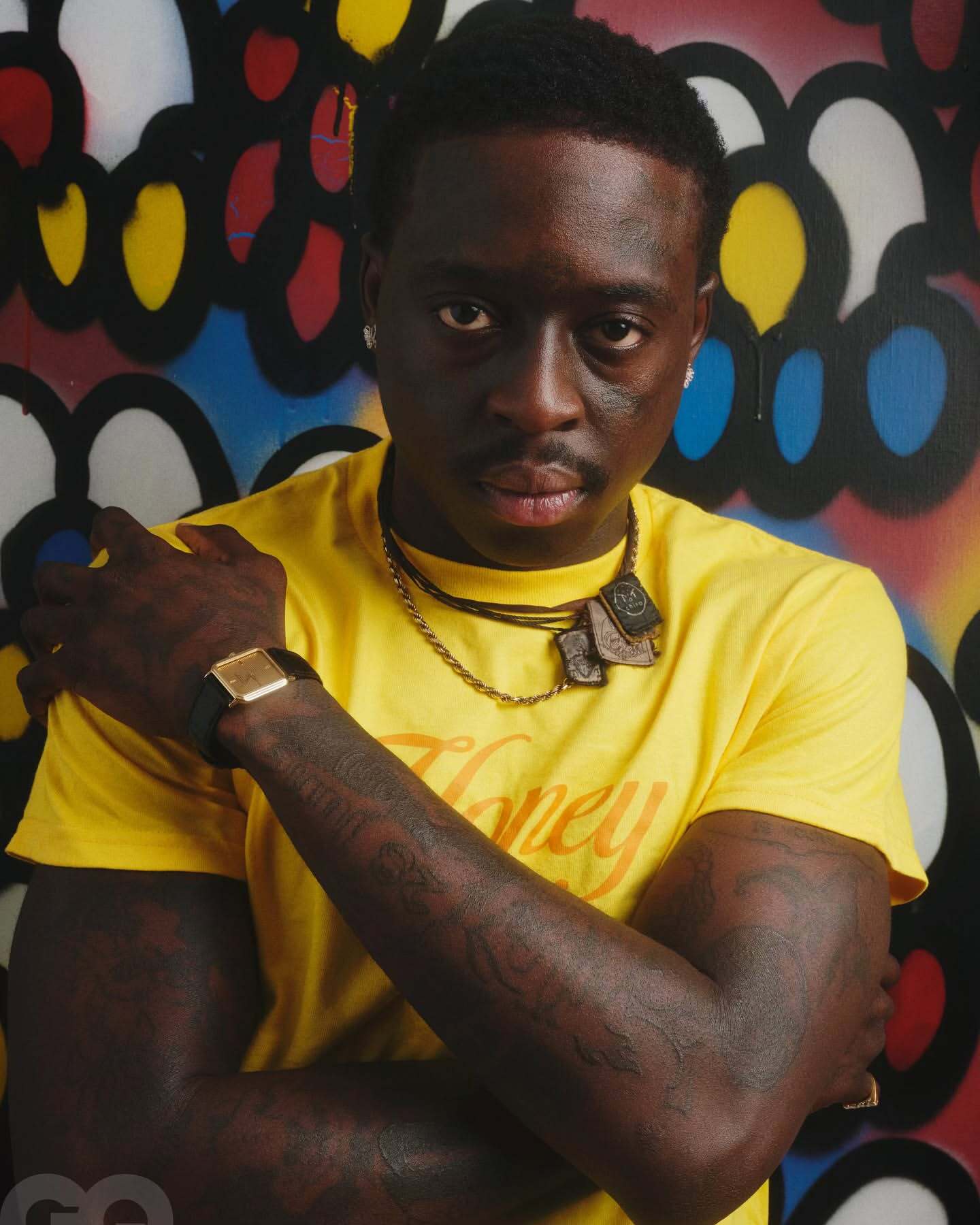 Portrait of an adult male with tattoos wearing a yellow T-shirt and jewelry, posing in front of a colorful graffiti-style backdrop.