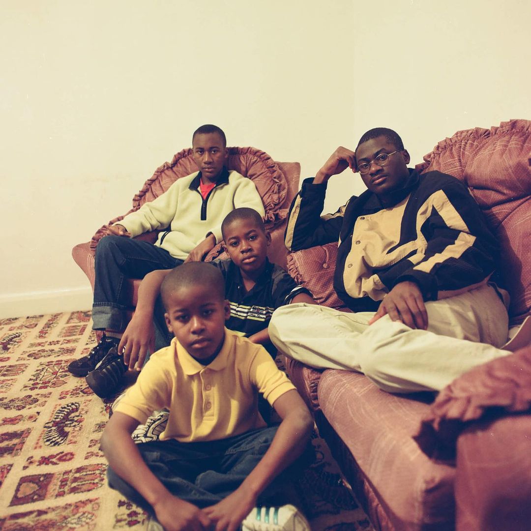 Group portrait of four African American males indoors: two adults and two children sit on floral sofas in casual clothes, looking at the camera.