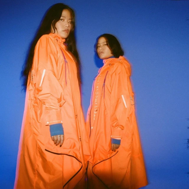 Two women stand in an indoor photo studio in bright orange raincoats against a blue background, captured in a medium shot.
