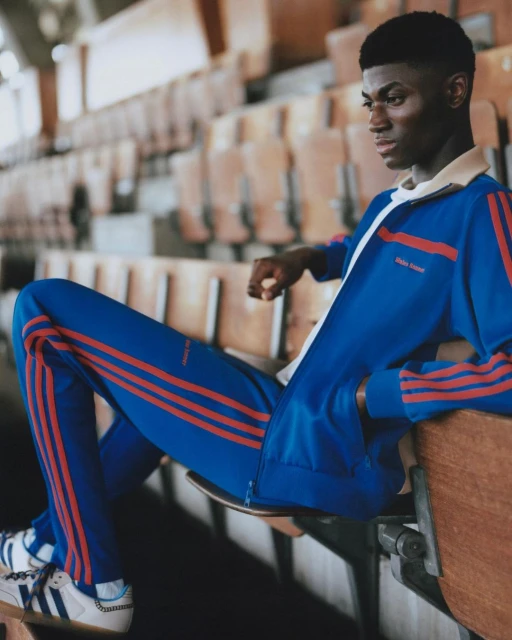 An athletic young Black man poses in a blue and orange track suit on wood bleachers in a sports stadium. He is looking off to the side.