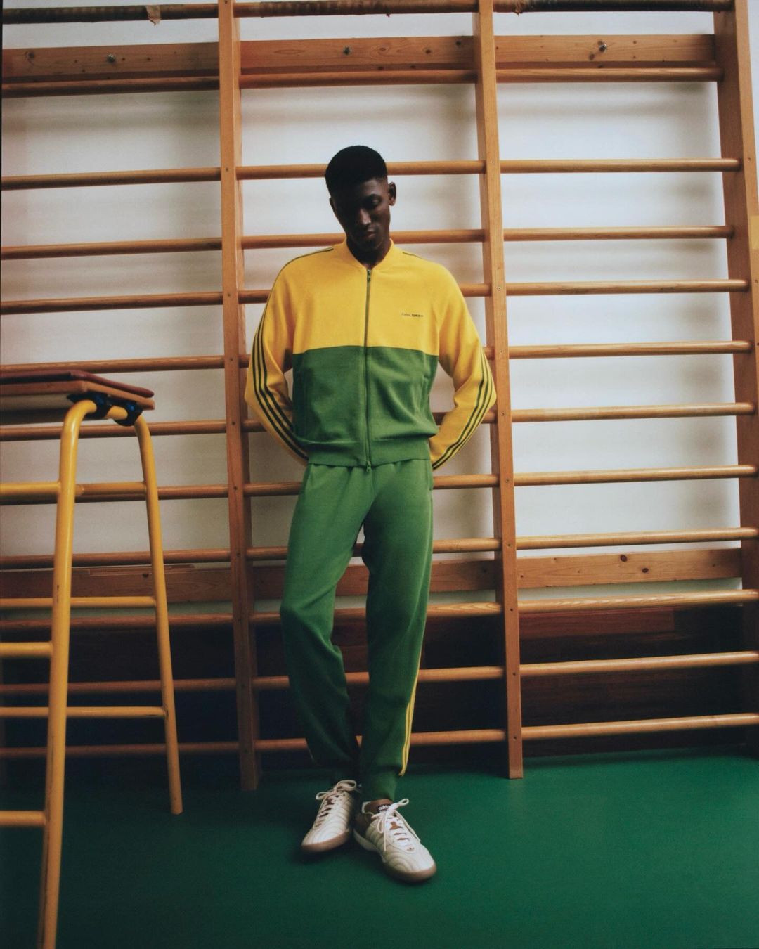 Man in retro-style tracksuit standing in a gym. The two-tone green and yellow tracksuit is vintage-inspired. He wears matching sneakers.