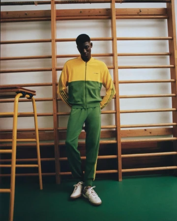 Man in retro-style tracksuit standing in a gym. The two-tone green and yellow tracksuit is vintage-inspired. He wears matching sneakers.