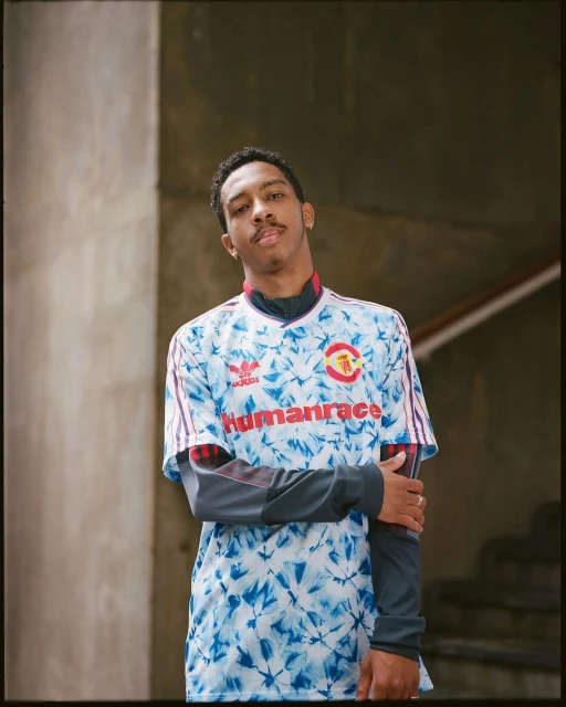 Man in a tie-dye soccer jersey with the word HUMANRACE across the chest stands in front of a staircase and concrete walls.