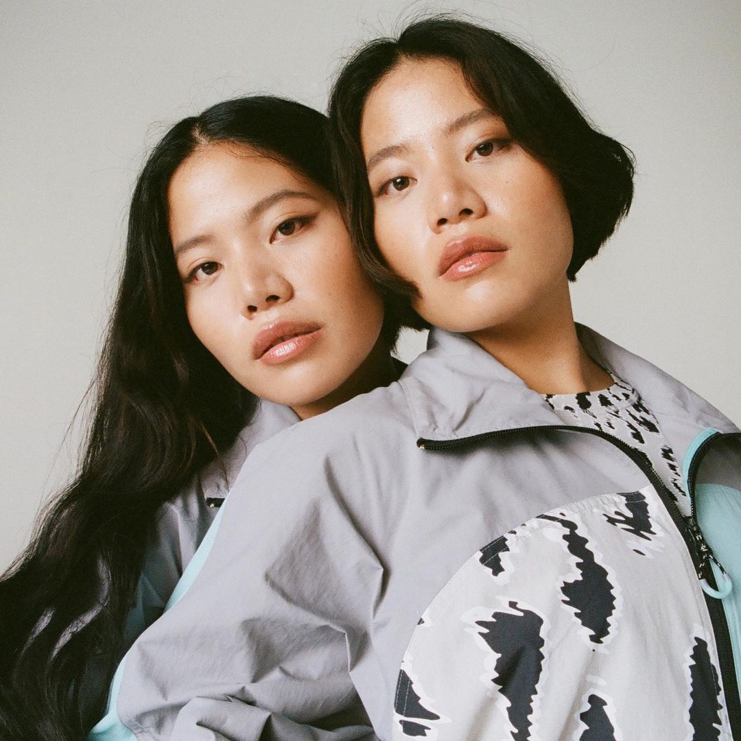 Portrait of two Asian women in grey jackets, one with long black hair and the other with short black hair, in a neutral-toned studio.