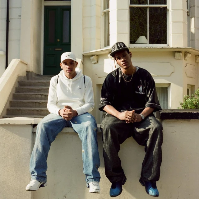 Two adult men sit outside a cream-colored building on the stoop in casual street fashion with a green door and white window frames.