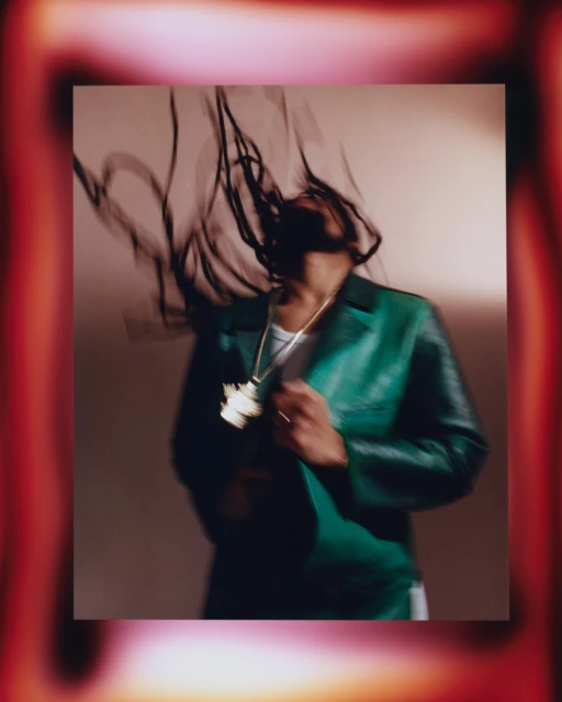 Portrait of a person with dreadlocks wearing a green leather jacket and gold chain necklace, with motion blur and soft lighting.