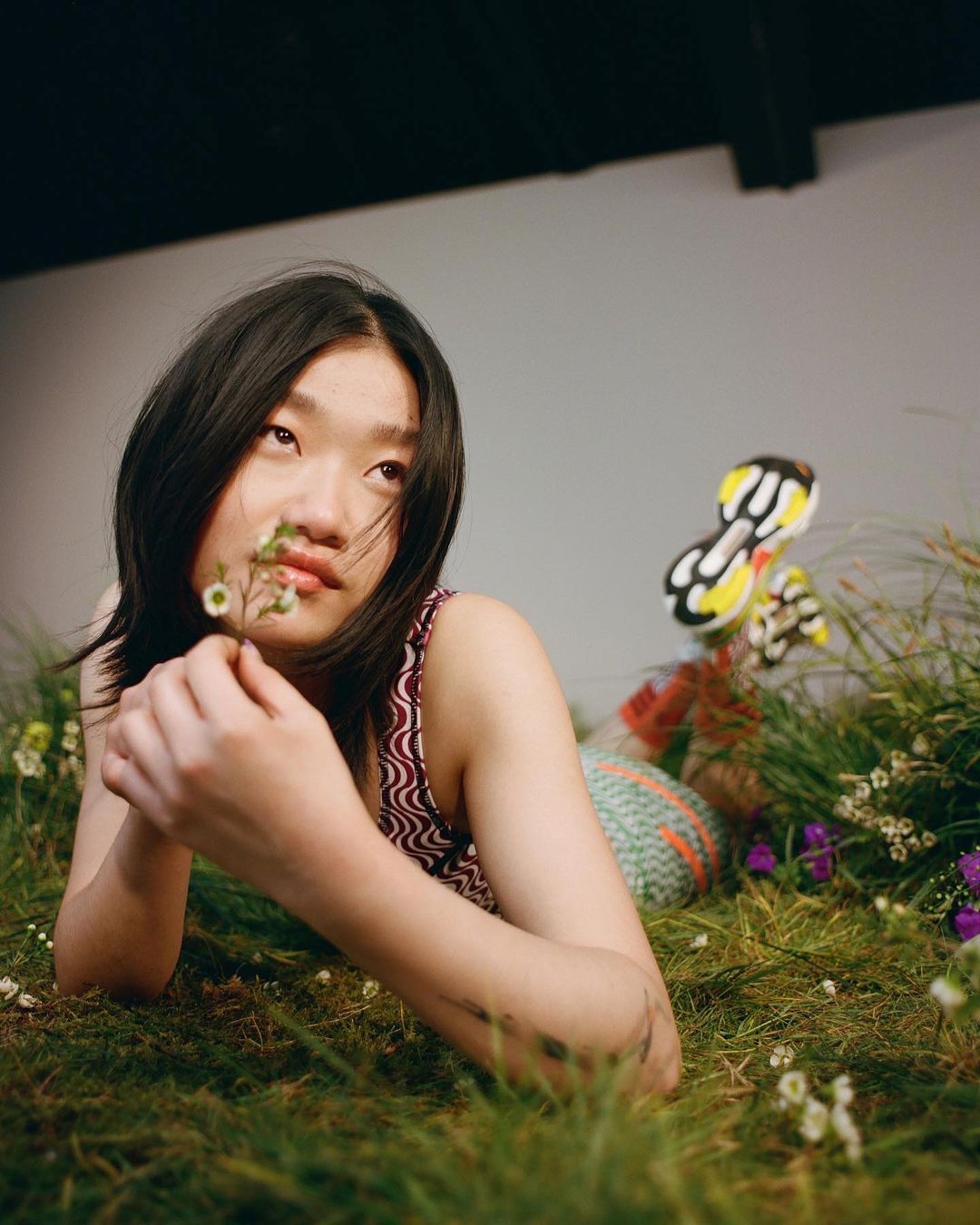 Young Asian woman lies in a field of green grass and small white flowers, holding a small sprig of flowers, looking upward.