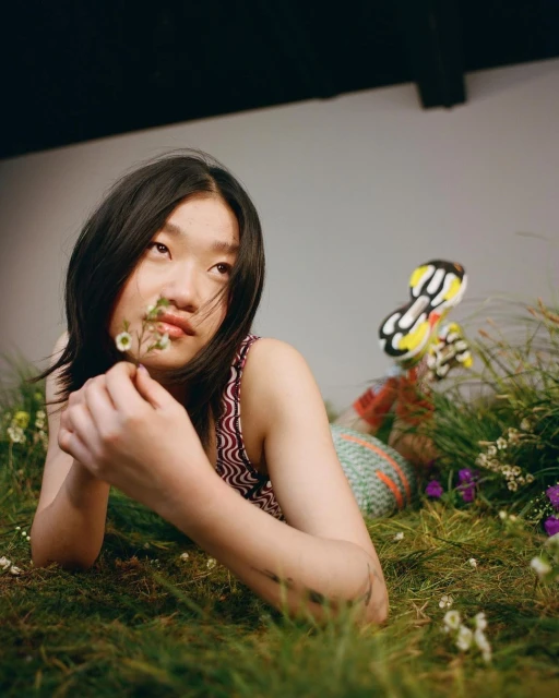 Young Asian woman lies in a field of green grass and small white flowers, holding a small sprig of flowers, looking upward.