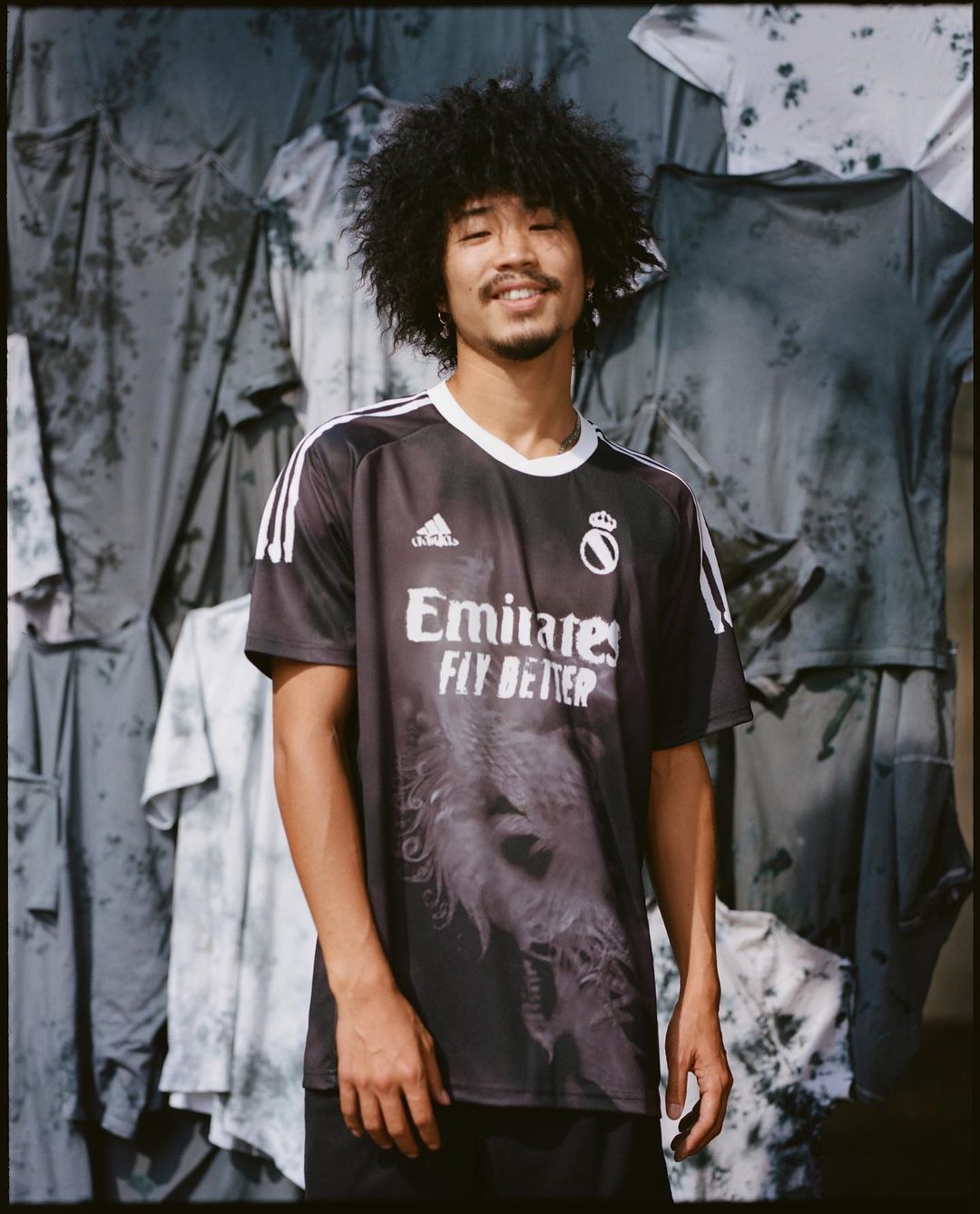Man in Real Madrid soccer jersey posing in front of tie-dye shirts; dark hair, smiling; sportswear; indoor lighting.