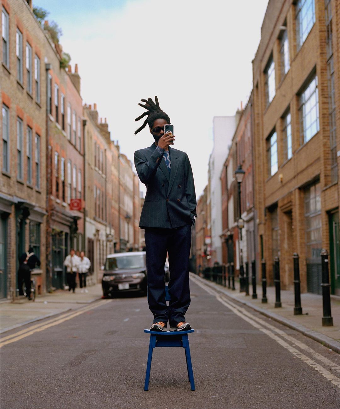 An adult male in a suit stands on a blue chair in the middle of a city street, holding a smartphone. Buildings line the street.