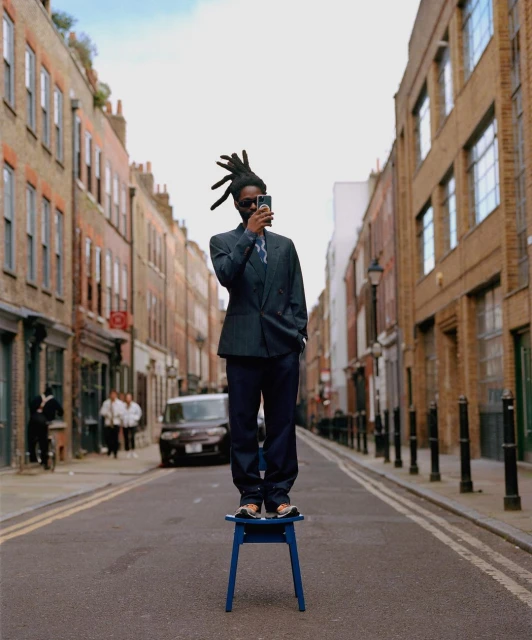 An adult male in a suit stands on a blue chair in the middle of a city street, holding a smartphone. Buildings line the street.