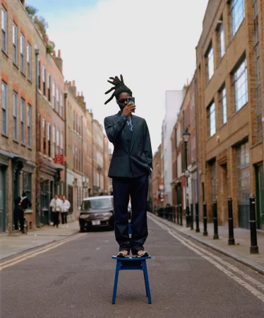 An adult male in a suit stands on a blue chair in the middle of a city street, holding a smartphone. Buildings line the street.