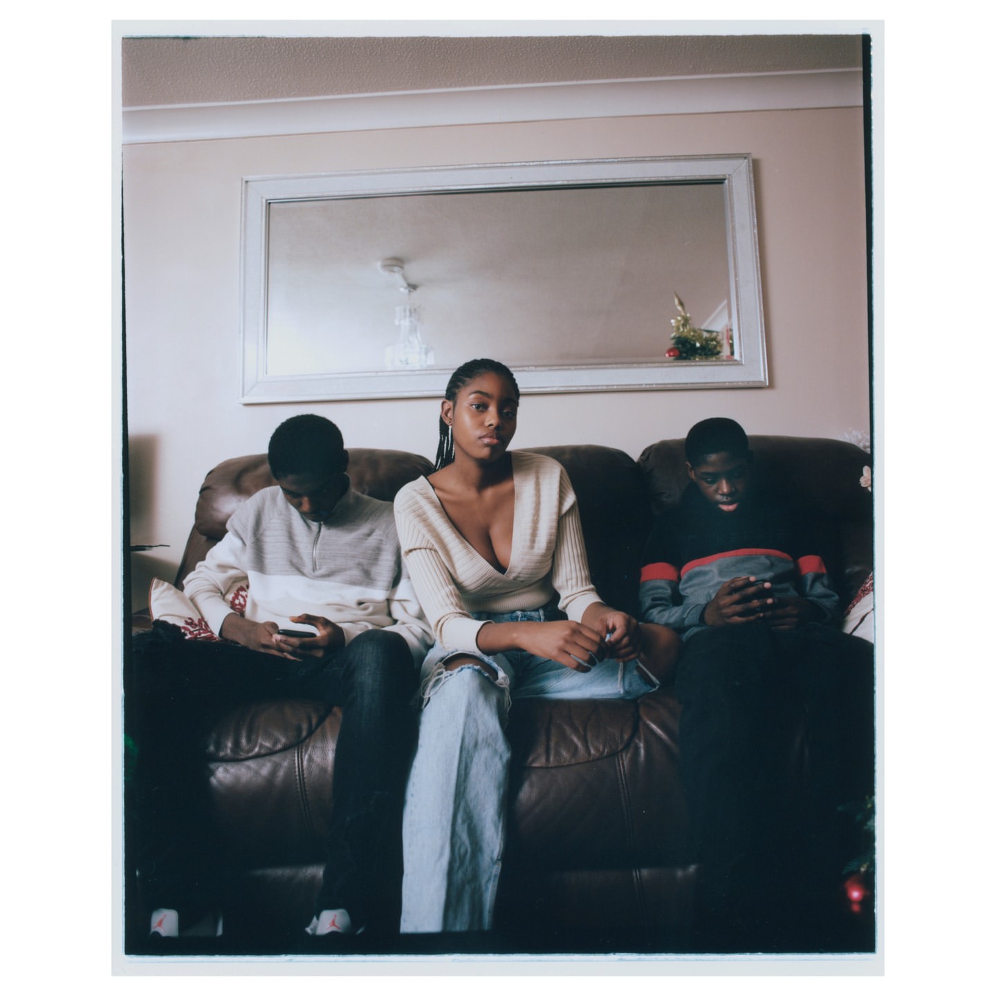 Three young adults (two males, one female) sit on a brown leather couch in a home interior. The woman is in the center wearing jeans.