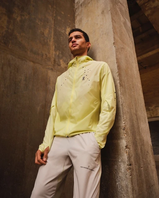 Athletic man in a yellow hooded jacket and grey pants standing against a textured concrete wall, looking up and to the left.