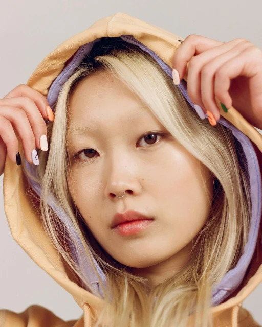 Portrait of an East Asian woman wearing a hooded sweatshirt, showcasing trendy multicolored nail art and natural makeup in a studio setting.
