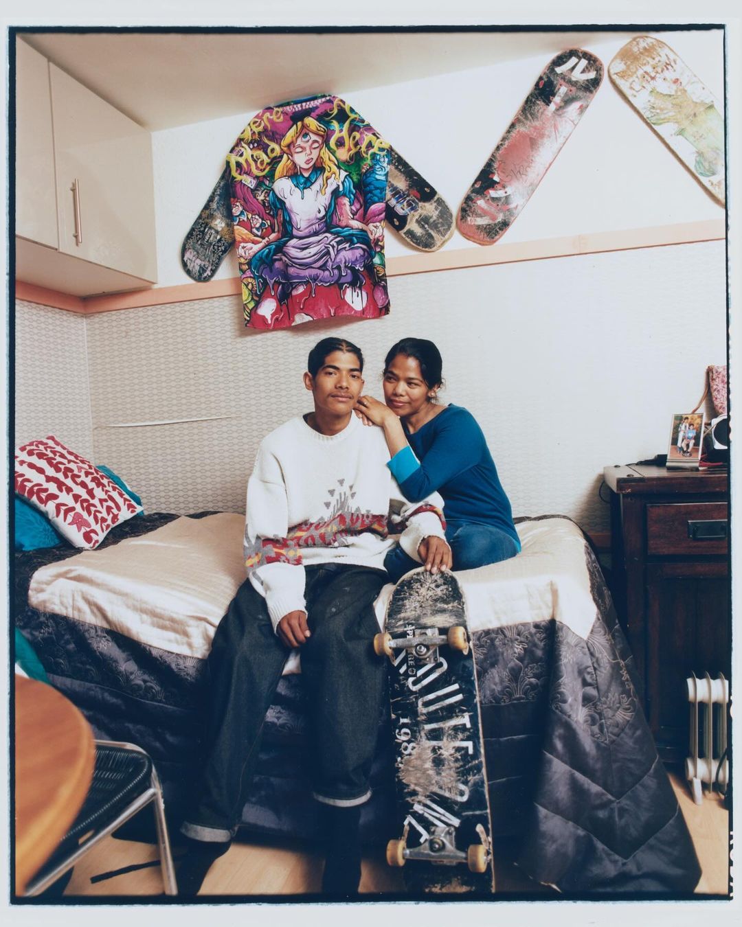Couple poses in indoor setting with skateboards and graphic art. The man sits with a skateboard, while a woman stands behind him.