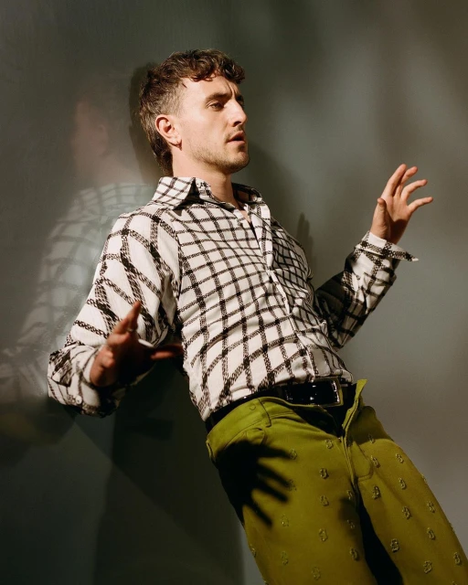 Adult man wearing a checkered shirt and patterned green pants leans against a wall in a studio setting with natural-looking light.