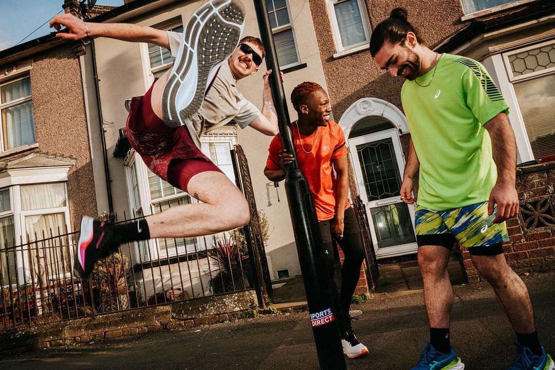 Three athletes wearing athletic gear and sneakers pose by a utility pole in front of townhouses on a sunny day, with one athlete jumping.