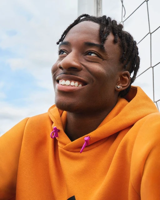 Close-up portrait of a smiling Black man with dreadlocks wearing an orange hoodie, looking upwards outdoors against a cloudy sky.